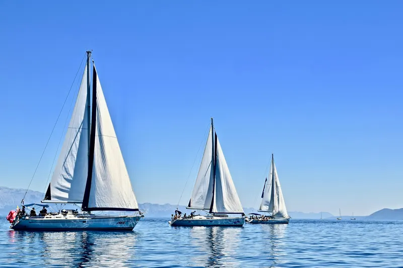 Tres veleros navegando en fila con el mar en calma y la costa montañosa al fondo.